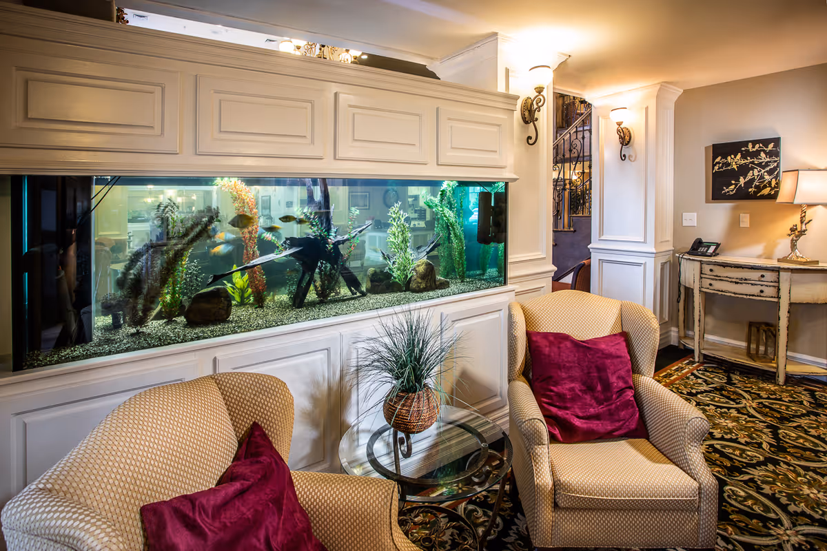 A cozy seating area in a senior living facility featuring two beige armchairs with red cushions, a glass-top side table with a potted plant, and a large built-in aquarium filled with fish and aquatic plants. The room has warm lighting, decorative wall sconces, a patterned carpet, and a small distressed wooden console table with a lamp and a phone.