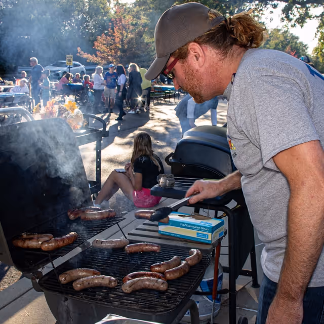 A man wearing a gray t-shirt and cap is grilling sausages on a large outdoor barbecue grill. Smoke rises from the grill as the man uses tongs to turn the sausages. In the background, people are gathered outdoors, some sitting and some standing, enjoying a sunny day with trees and autumn foliage visible.