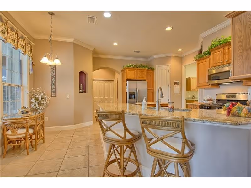 A bright kitchen area with a granite countertop island featuring two wooden bar stools. The kitchen has wooden cabinets, a stainless steel refrigerator, and a stove. To the left, there is a small dining nook with a round table and two chairs near large windows with floral valances. The floor is tiled, and the walls are painted beige with recessed lighting overhead.