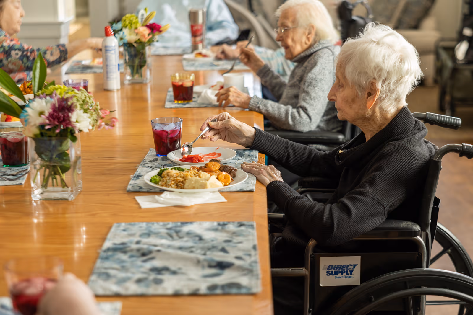 Elderly residents sitting at a long wooden dining table in a senior living facility, eating a meal. The table is decorated with vases of flowers and placemats. One woman in a wheelchair is in the foreground, holding a spoon and eating from a plate of food. Other residents are seated along the table, engaged in eating and conversation.