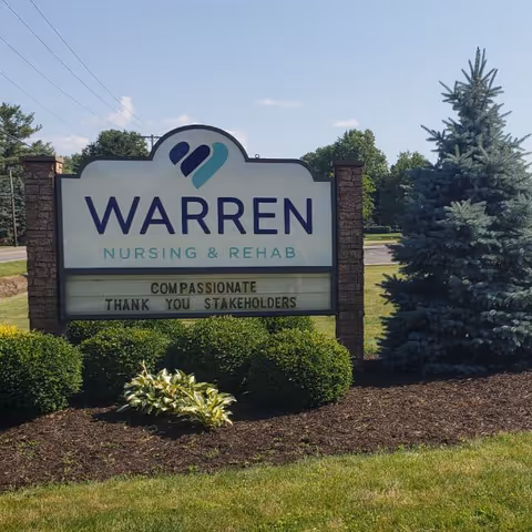 Outdoor sign reading 'Warren Nursing & Rehab' on a landscaped lawn with bushes and a pine tree under a blue sky.