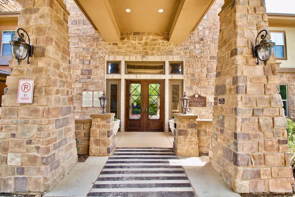 Entrance to a building with large stone pillars on either side, a striped mat on the ground, and double wooden doors with glass panels. There are wall-mounted lantern-style lights on the pillars and signs indicating no parking and assisted living.