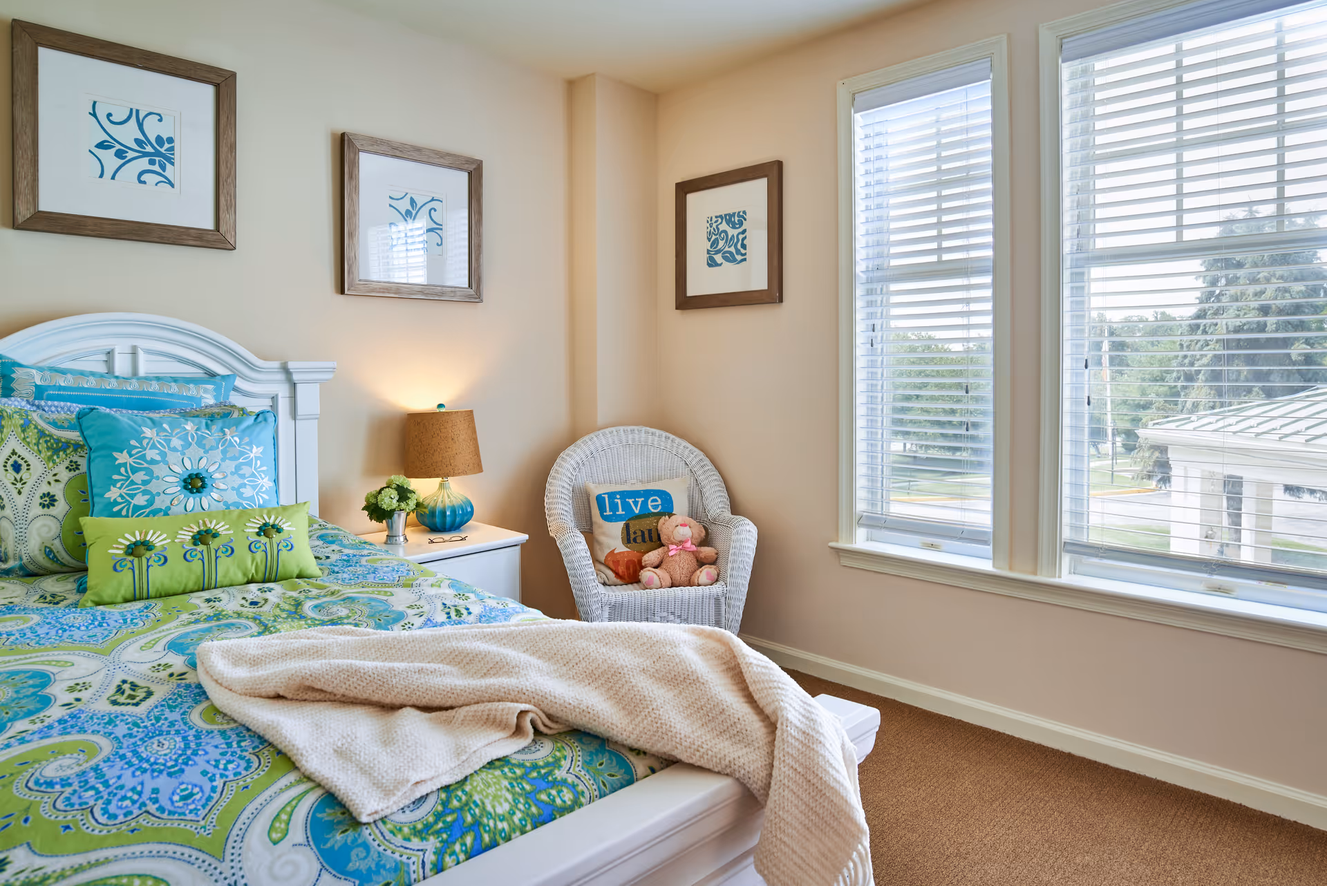 Sunlit bedroom with a white bed dressed in green and blue patterned linens, a nightstand with a lamp, a wicker chair holding a pillow and stuffed toys, and large windows with blinds.