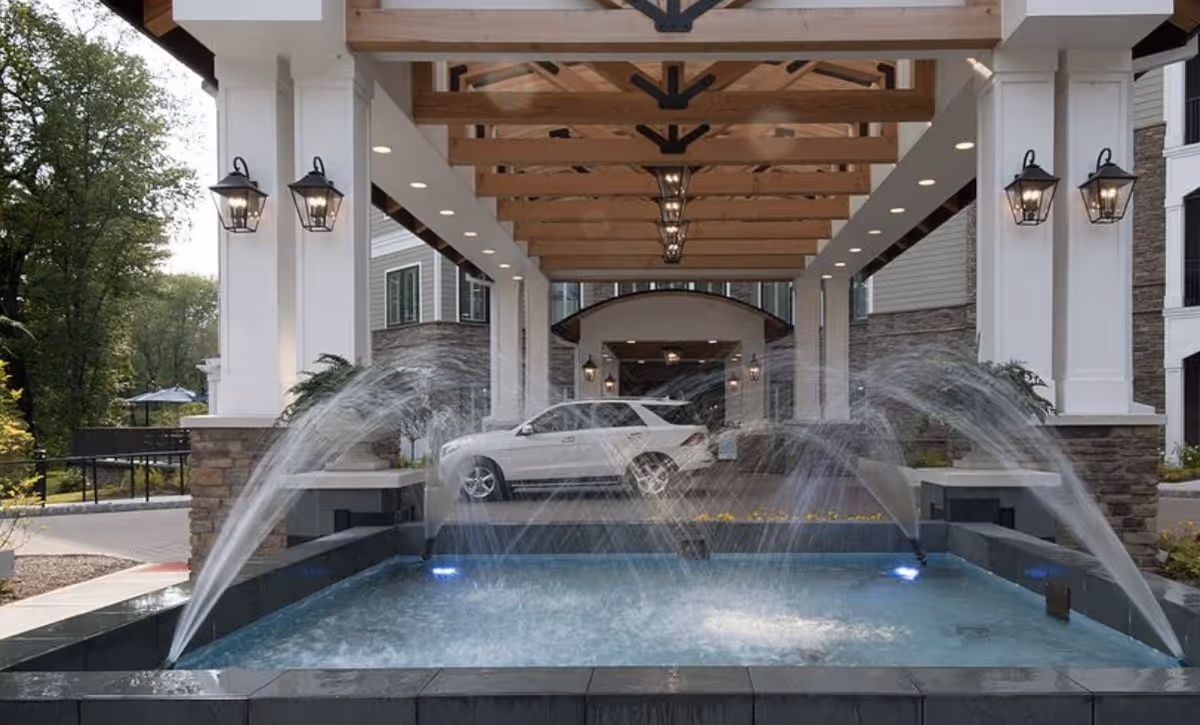 Covered entrance of a senior living facility with a white SUV parked under a wooden beam canopy. In front of the entrance is a rectangular water fountain with multiple water jets spraying water upwards. The building exterior features stone and white pillars with black lantern-style lights mounted on the pillars.