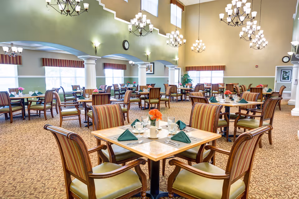 Spacious dining room with multiple set tables and striped chairs under chandeliers in a senior living facility.