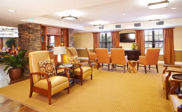 Spacious senior living lounge with upholstered armchairs, a central meeting table, TV on the wall, and large windows.