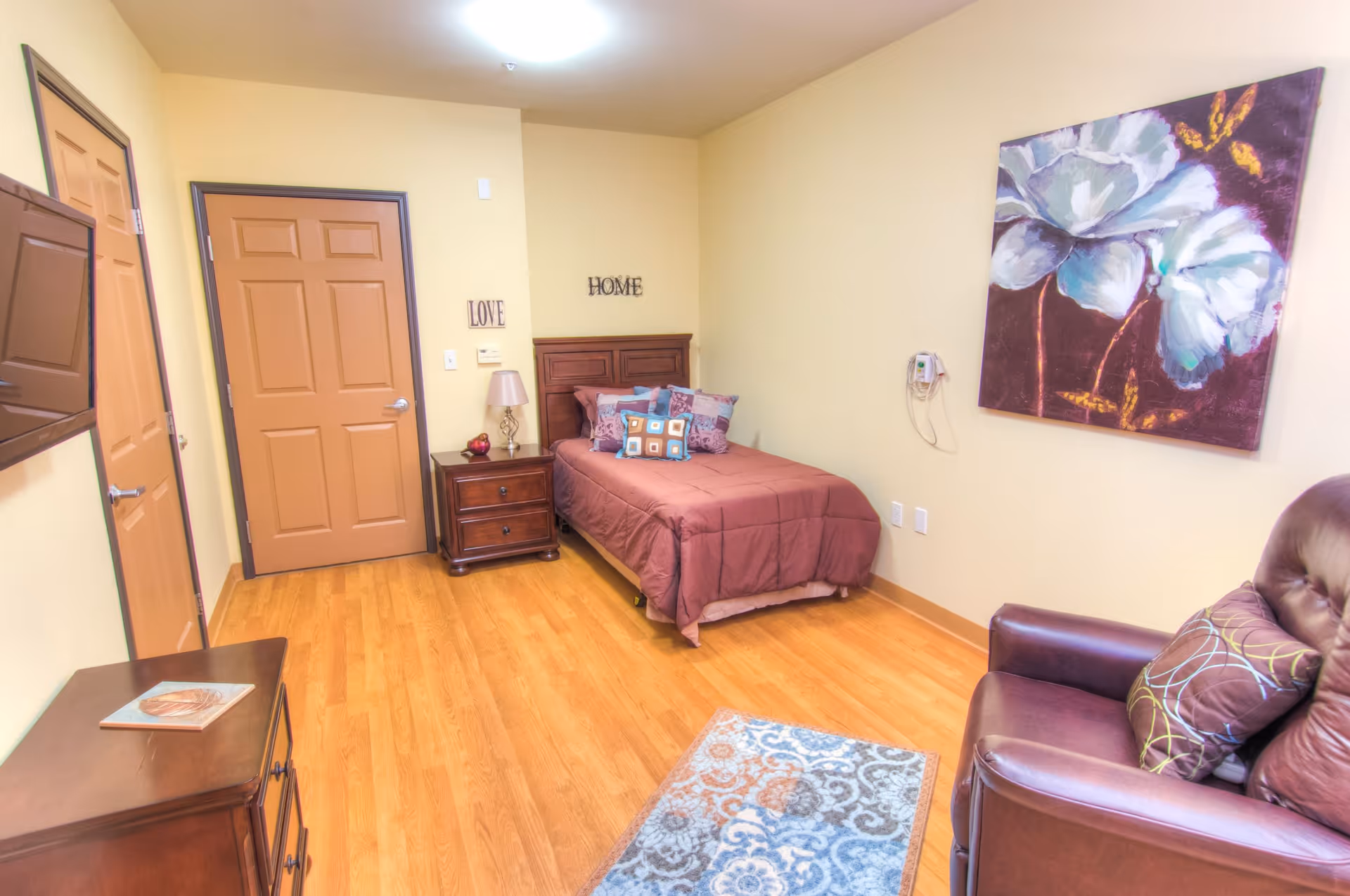 A cozy bedroom with a single bed covered in a brown comforter and multiple decorative pillows. There is a wooden nightstand with a lamp and a small decorative item next to the bed. The walls are painted light yellow, with the words 'LOVE' and 'HOME' hanging above the bed. A large floral painting is mounted on the wall to the right. A brown leather armchair with a patterned pillow is positioned near the painting. The floor is wooden with a small patterned rug in front of the chair. Two closed doors and a wall-mounted TV are visible on the left side of the room.