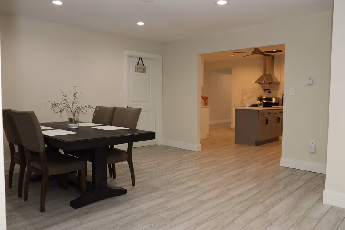 Interior view of a dining area with a dark wooden table and four upholstered chairs. A small vase with greenery is placed on the table. The room has light-colored walls and wood-look tile flooring. In the background, there is an open doorway leading to a kitchen with gray cabinets, a stainless steel range hood, and a stove.