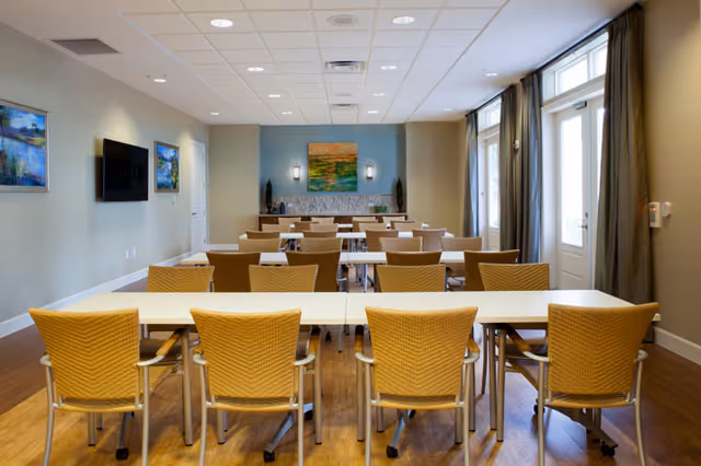 A bright and clean dining room with multiple tables and wicker chairs arranged in rows. The room features wood flooring, a wall-mounted TV, framed artwork on the walls, large windows with curtains, and a door leading outside. The far wall has a decorative fireplace with two small plants on either side and a colorful painting above it.