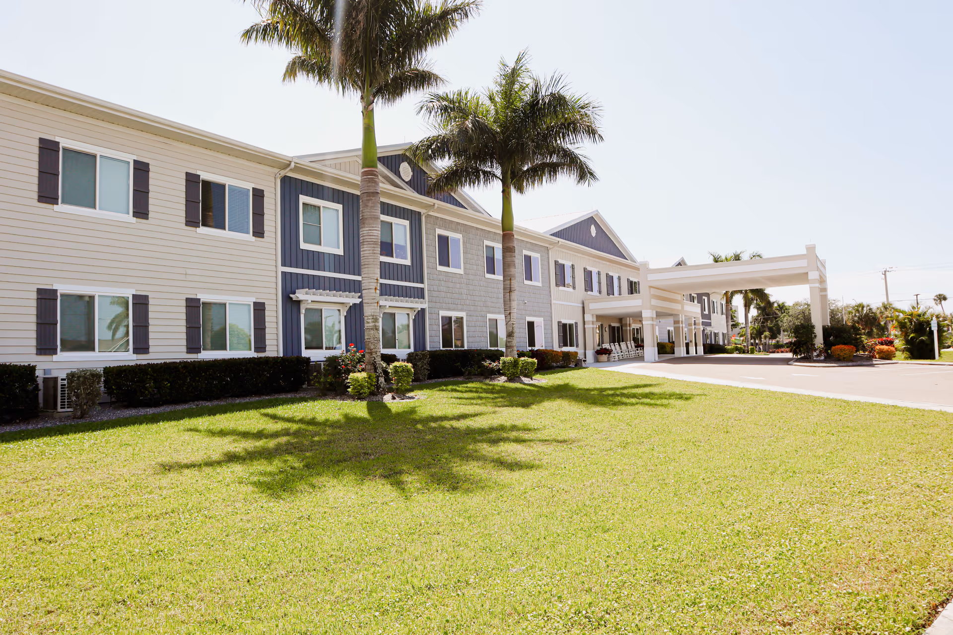 Exterior view of a two-story senior living facility building with beige and blue siding, multiple windows, palm trees, and a covered entrance driveway. The foreground shows a well-maintained green lawn under a clear sky.
