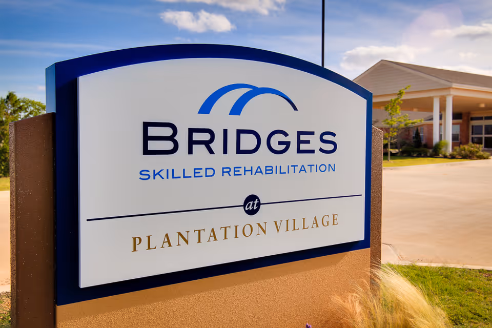 Outdoor sign for Bridges Skilled Rehabilitation at Plantation Village Nursing Center with a building and driveway in the background under a partly cloudy sky.
