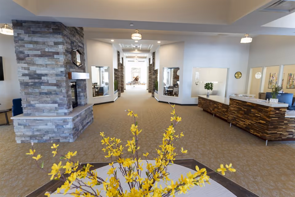 Spacious senior living facility lobby with a stone fireplace, reception desk, and a yellow floral centerpiece in the foreground.