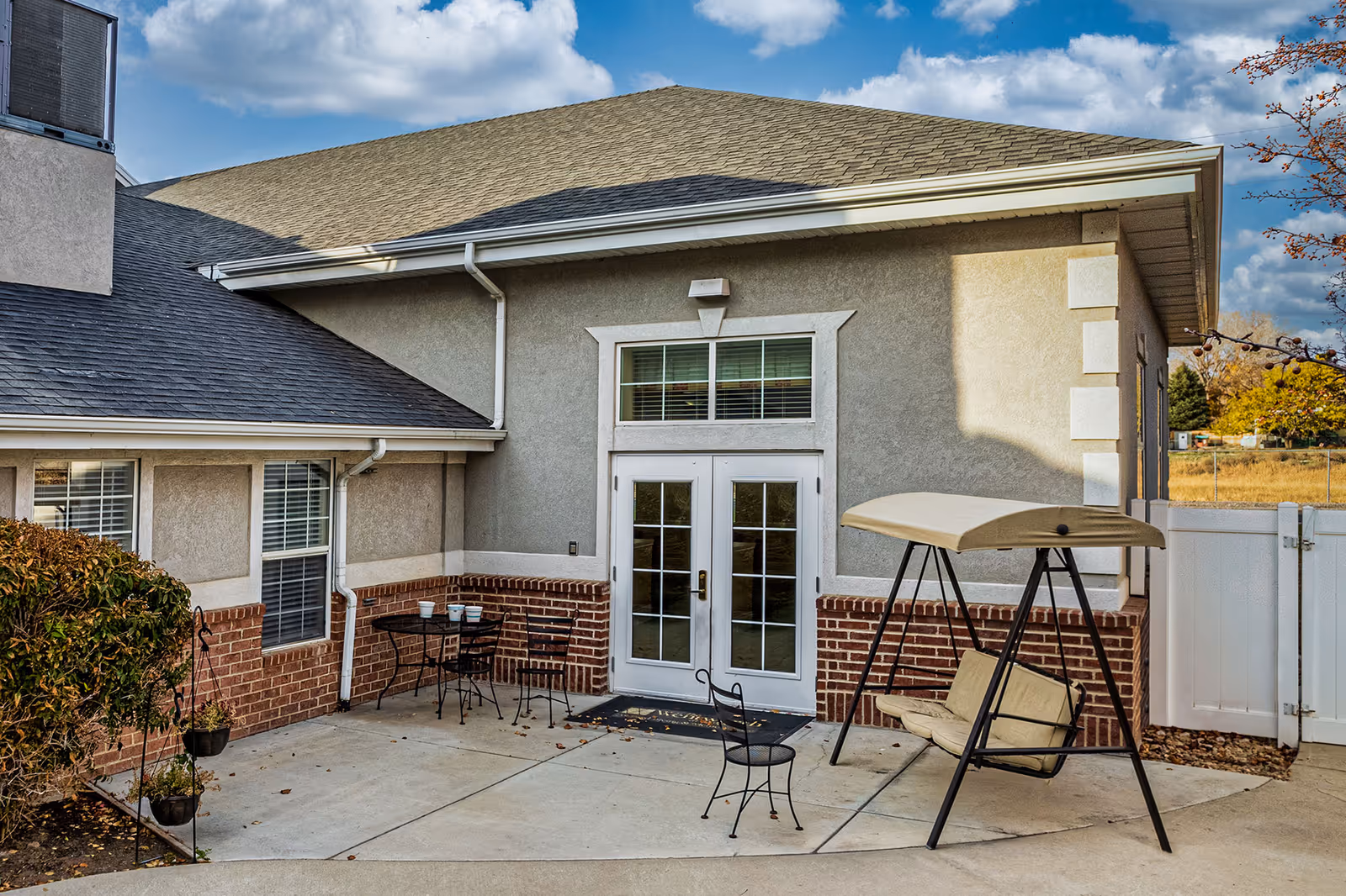 Outdoor patio area at Valencia at Draper featuring a beige cushioned swing with a canopy, a small black metal table with two chairs, and a single black metal chair. The patio is adjacent to a building with beige and brick exterior walls, white-framed double glass doors, and windows with white blinds. The sky is partly cloudy.