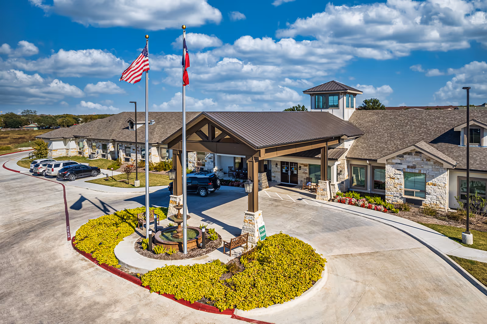 Exterior view of a senior living facility with a covered entrance, two flagpoles displaying the American and Texas flags, landscaped greenery, parked cars, and a partly cloudy blue sky.