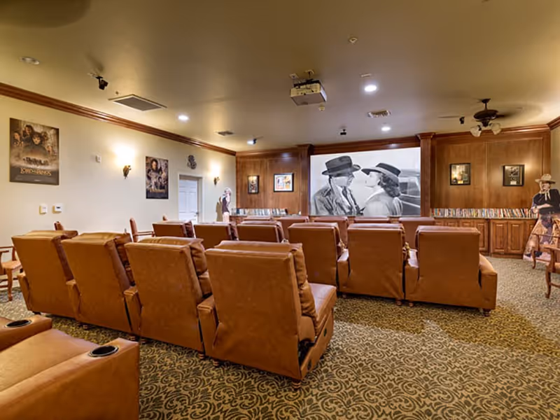 A cozy movie theater room with multiple rows of brown leather recliner chairs facing a large screen showing a black and white movie scene. The room has patterned carpet, wood-paneled walls, movie posters, and a ceiling-mounted projector. There are also life-sized cutouts of movie characters near the walls.