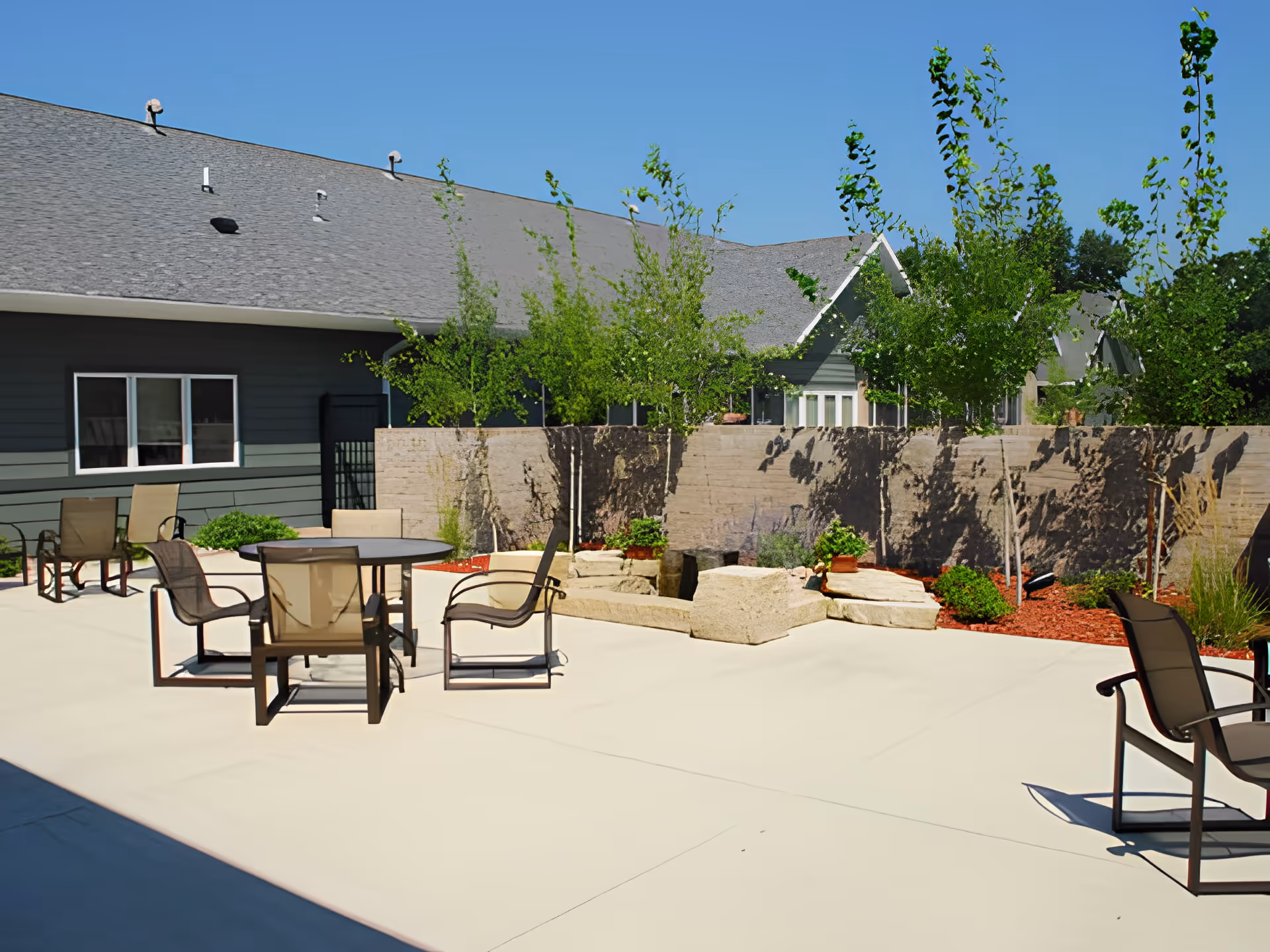 Outdoor patio area at Friendship Haven with several chairs and round tables on a concrete surface, surrounded by a low stone wall and young trees, under a clear blue sky.