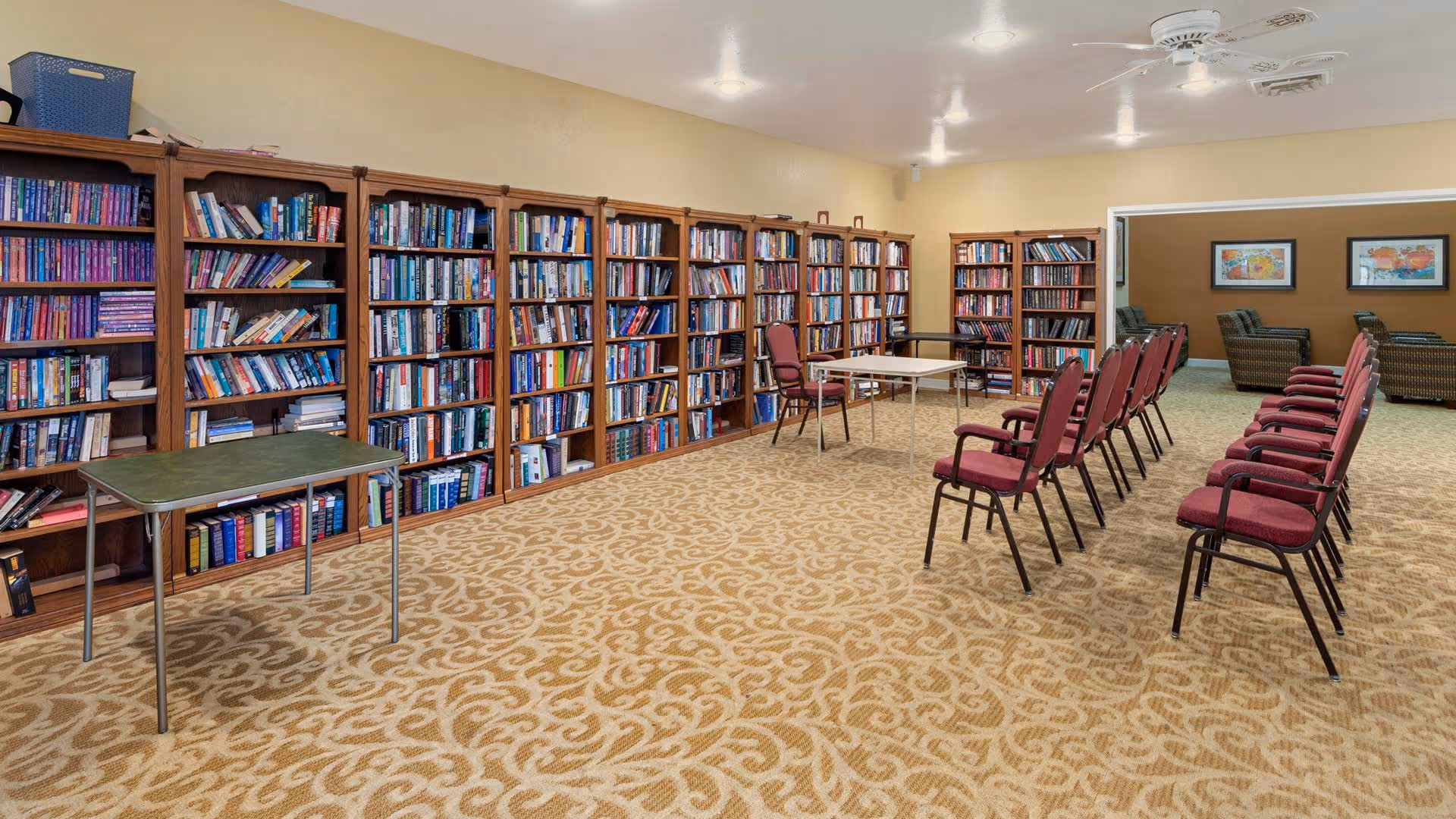 Spacious interior library/multipurpose room with wall-length bookshelves, rows of maroon chairs, and folding tables on patterned carpet.