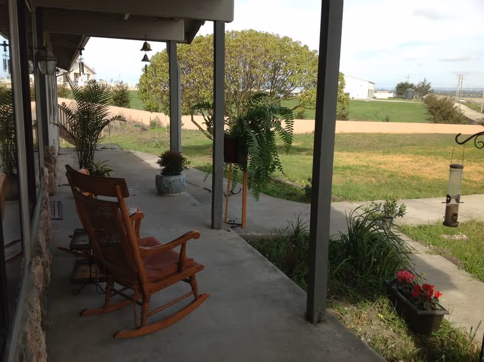 Covered outdoor patio area with a wooden rocking chair, potted plants, and a bird feeder hanging from a metal hook. The patio overlooks a grassy yard with trees and a distant building under a partly cloudy sky.