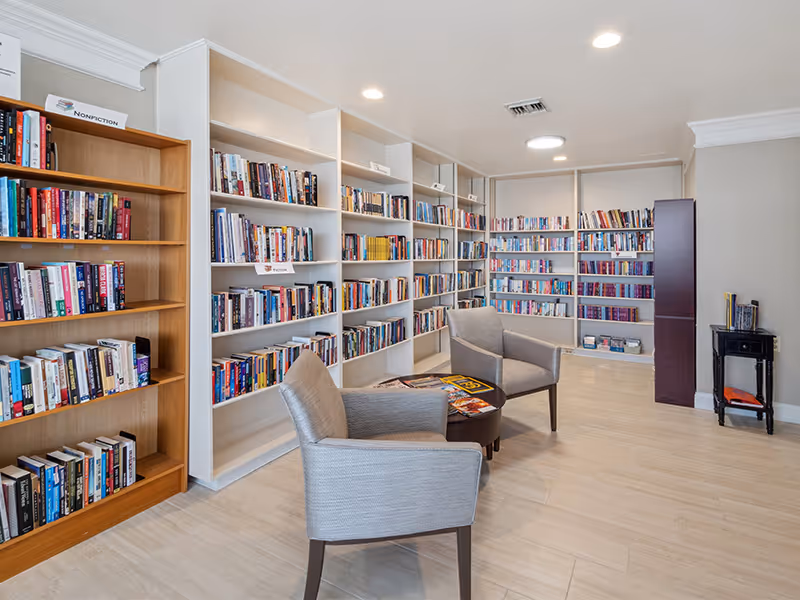 A well-lit library room with multiple white and wooden bookshelves filled with books. Two gray upholstered chairs are positioned around a small round table with magazines on it. The floor is light-colored wood, and the ceiling has recessed lighting.