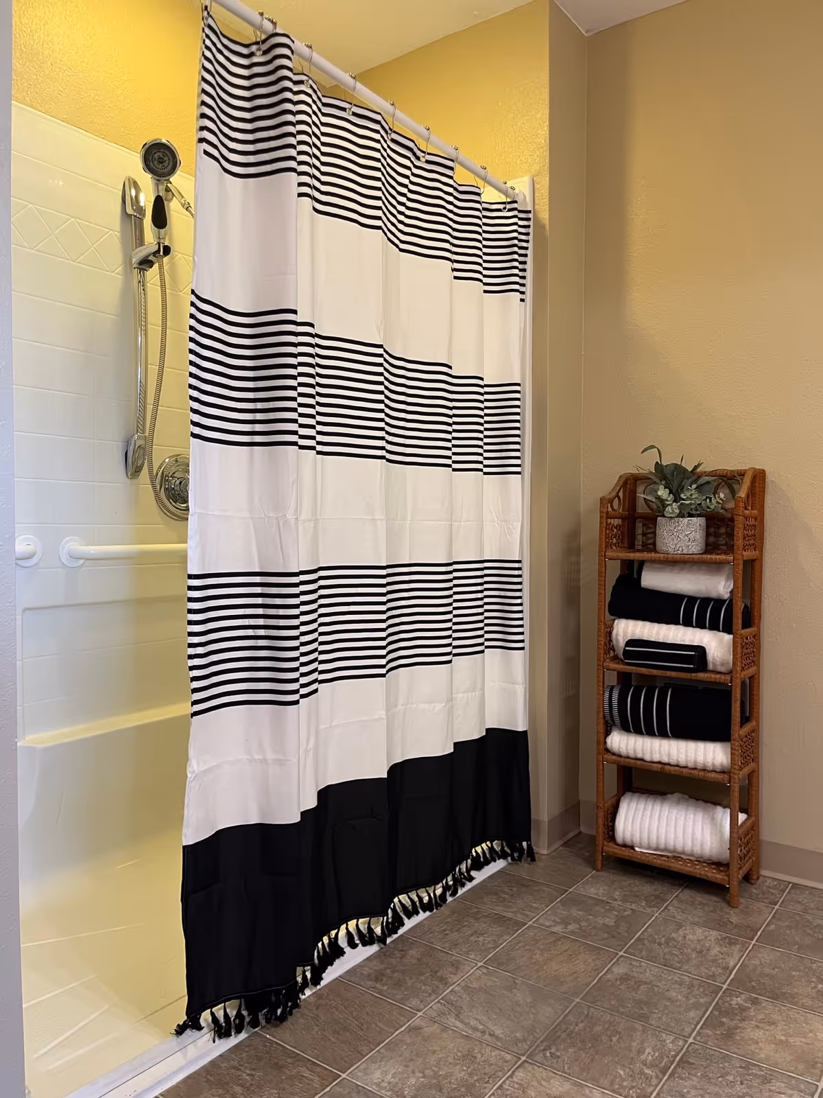 Bathroom with a white and black striped shower curtain partially covering a shower area with a handheld showerhead. To the right, there is a wooden shelf holding neatly folded black and white towels and a small potted plant on top. The floor is tiled and the walls are painted beige and yellow.