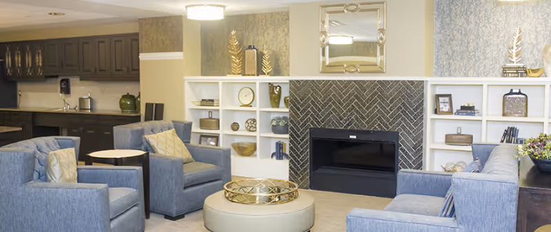 A cozy living room area in Inniswood Village featuring blue upholstered armchairs and sofas arranged around a round ottoman with a decorative tray. Behind the seating area is a fireplace with a herringbone tile surround, flanked by white built-in shelves holding various decorative items. To the left, there is a kitchenette with dark cabinetry and a small round table with chairs.