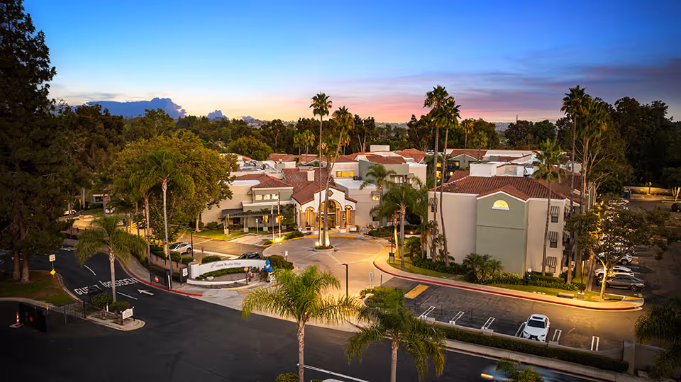 Aerial view of a senior living facility at dusk with Mediterranean-style buildings, palm trees, and a circular driveway entrance surrounded by landscaped greenery and parking spaces.