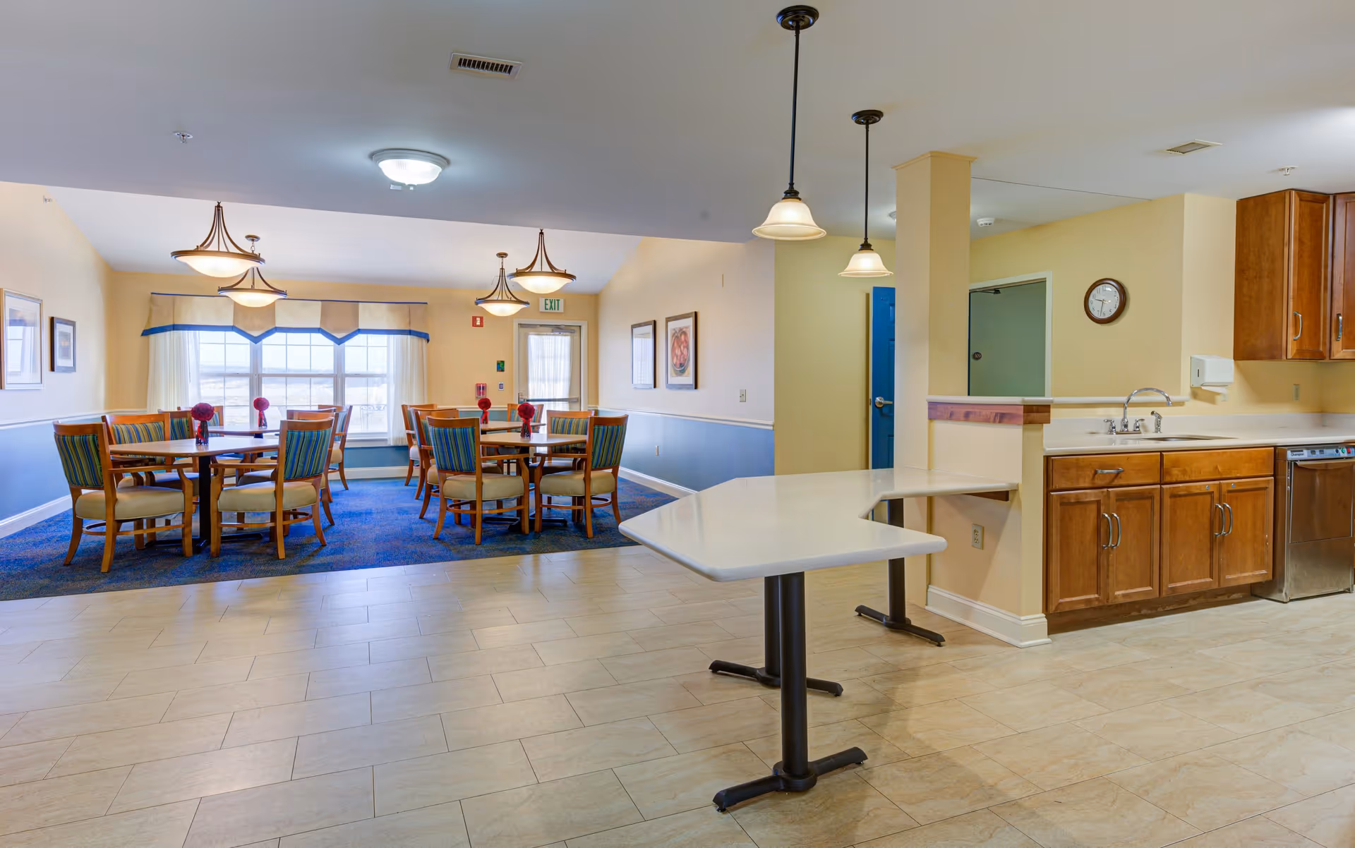 A bright dining area in a senior living facility with several wooden tables and chairs arranged on a blue carpet near large windows with white curtains. The foreground shows a kitchen counter with a sink, wooden cabinets, and hanging pendant lights above. The walls are painted in soft yellow and blue tones, and framed artwork is visible on the walls.