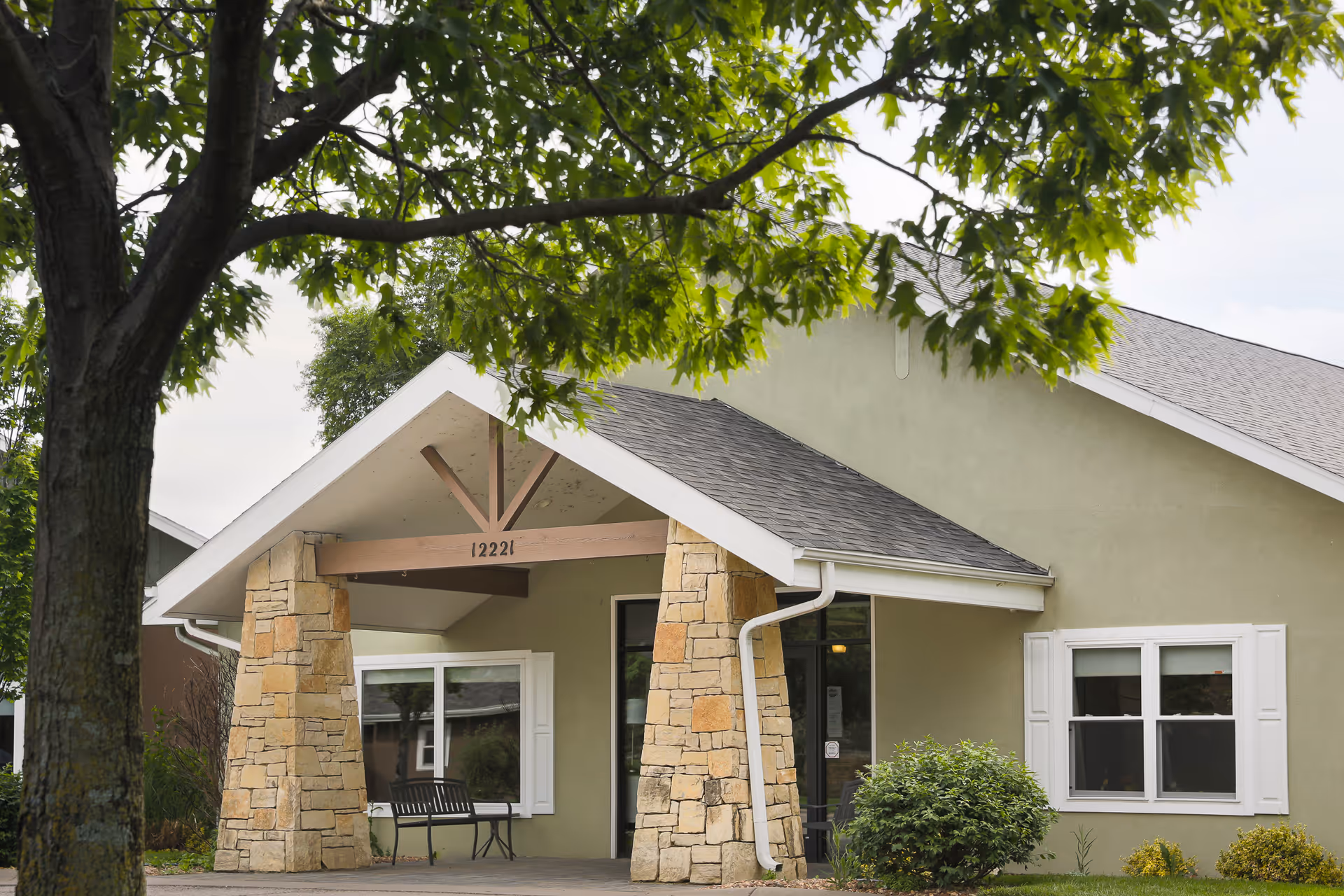 Front entrance of a single-story assisted living building with a covered porch supported by stone pillars, a bench, and a tree in the foreground.