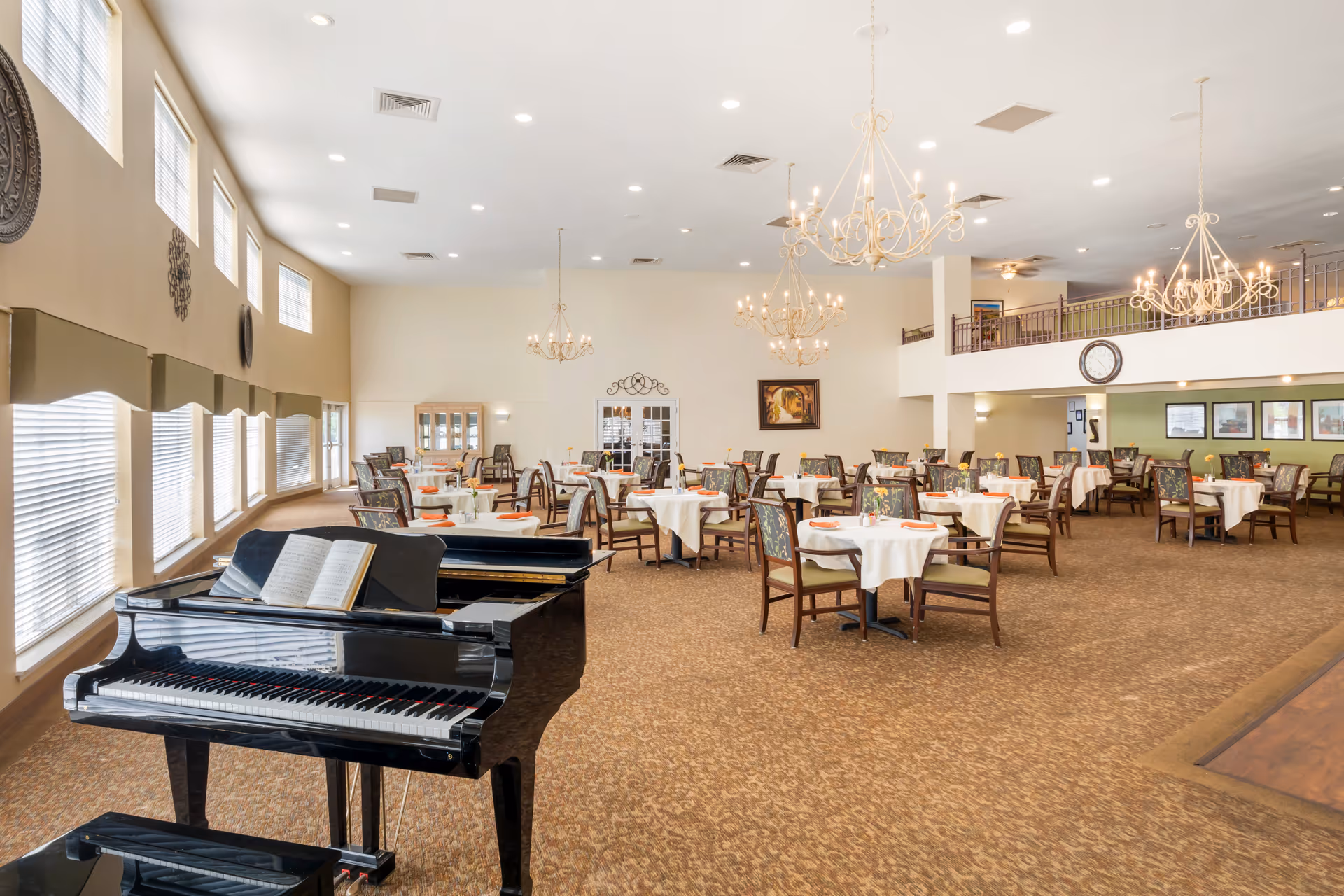 Spacious dining room with multiple round tables covered with white tablecloths and set with plates and napkins. The room features a grand piano with an open music book near large windows with blinds. Elegant chandeliers hang from the ceiling, and there is a mezzanine level with railings overlooking the dining area.