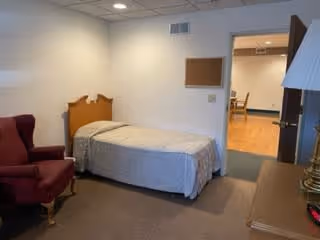 A simple bedroom in an assisted living facility featuring a single bed with a wooden headboard, a maroon armchair, a wooden dresser with a lamp, and a view into an adjacent room with wooden flooring and chairs.