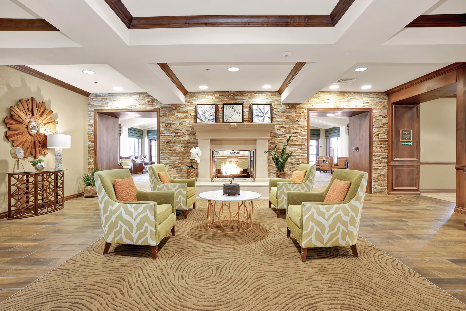 A cozy living room area with four green and white patterned armchairs arranged around a round coffee table on a textured beige rug. The room features a stone fireplace with a fire burning, flanked by plants and decorative artwork above the mantel. The walls have a mix of stone and wood paneling, and the ceiling has wooden beams. There are doorways leading to other rooms with natural light coming through windows.