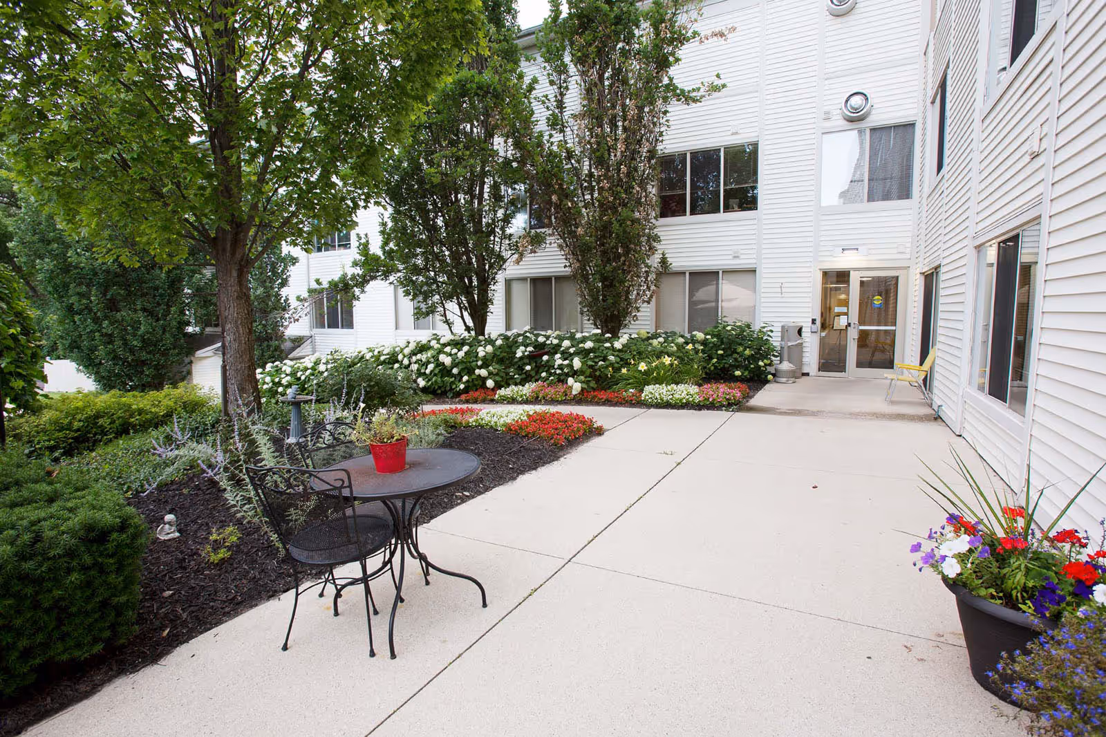 Outdoor patio area at Independence Village of Plymouth featuring a small round metal table with two chairs, surrounded by lush greenery, colorful flower beds, and trees. The white exterior of the building with multiple windows and a glass door entrance is visible in the background.