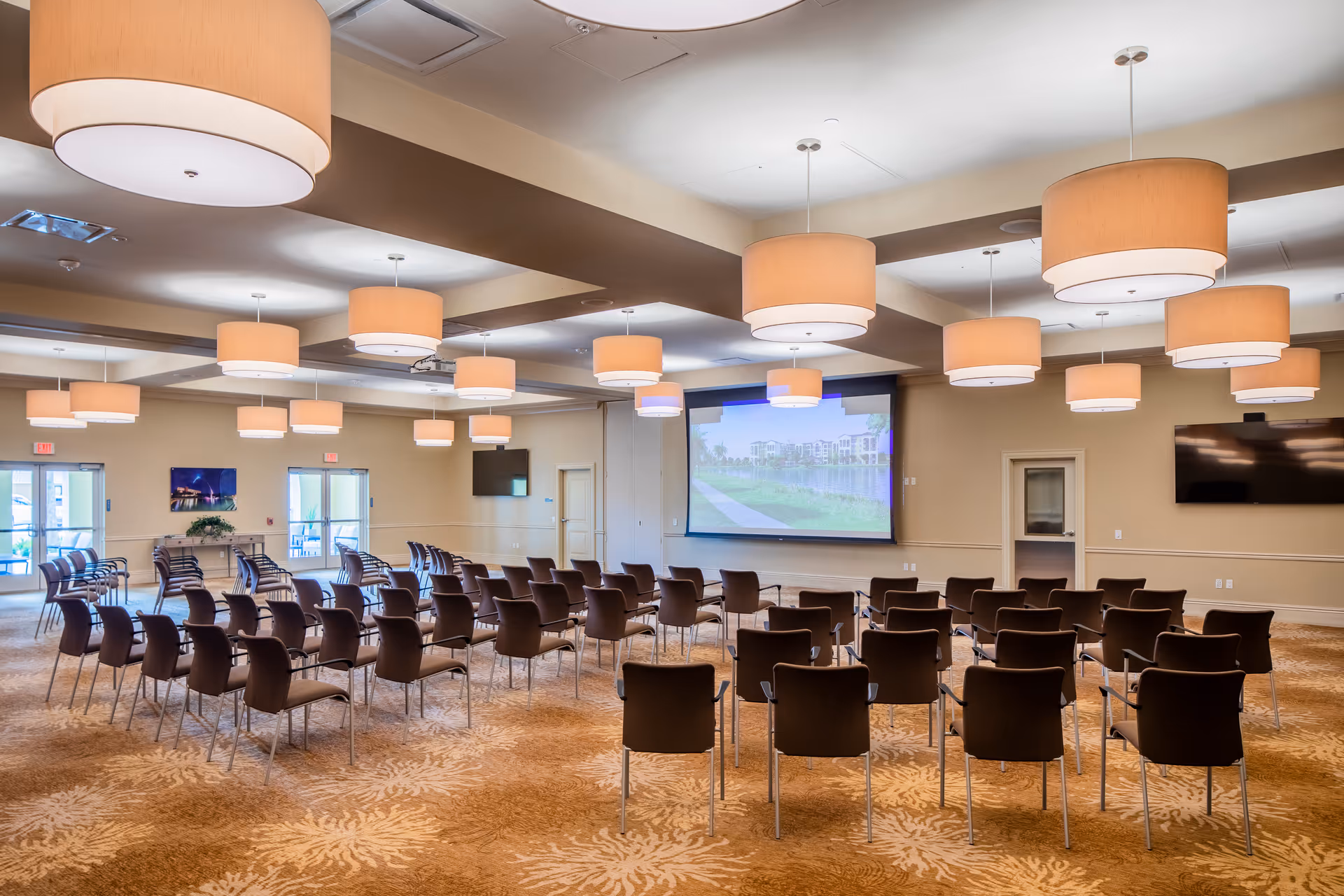 A large conference or meeting room with rows of black chairs arranged facing a large projection screen displaying an outdoor scene. The room has beige walls, multiple large round ceiling lights, and two flat-screen TVs mounted on the walls. There are double glass doors at the back of the room allowing natural light in.