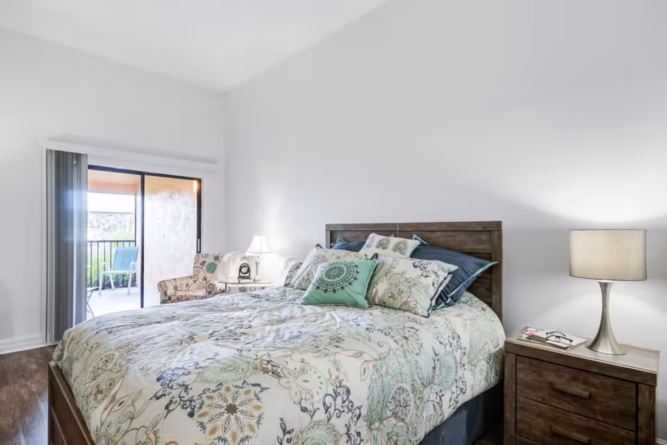 Bright bedroom with a patterned bed, wooden headboard, nightstands and a sliding glass door to a balcony.