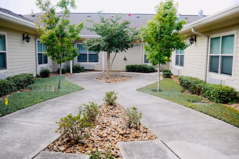 Outdoor courtyard area in a senior living facility with concrete walkways, small trees, bushes, and landscaping rocks. The courtyard is surrounded by single-story beige buildings with multiple windows.
