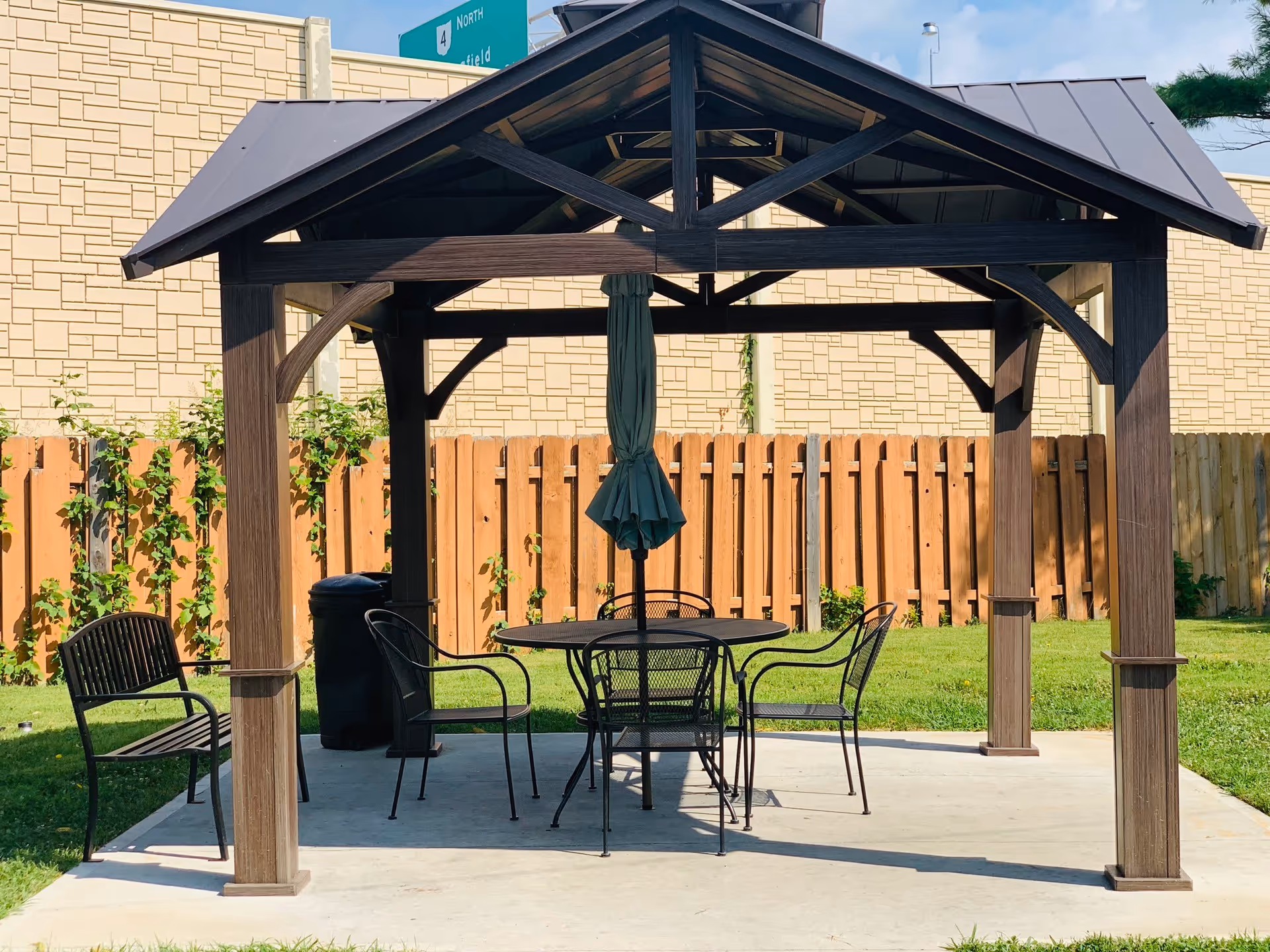 Outdoor seating area with a wooden gazebo structure covering a round metal table and four metal chairs, with a folded green umbrella in the center. There is a wooden bench to the left and a trash can behind the table. The area is surrounded by a wooden fence and green grass, with a beige wall and a road sign visible in the background.