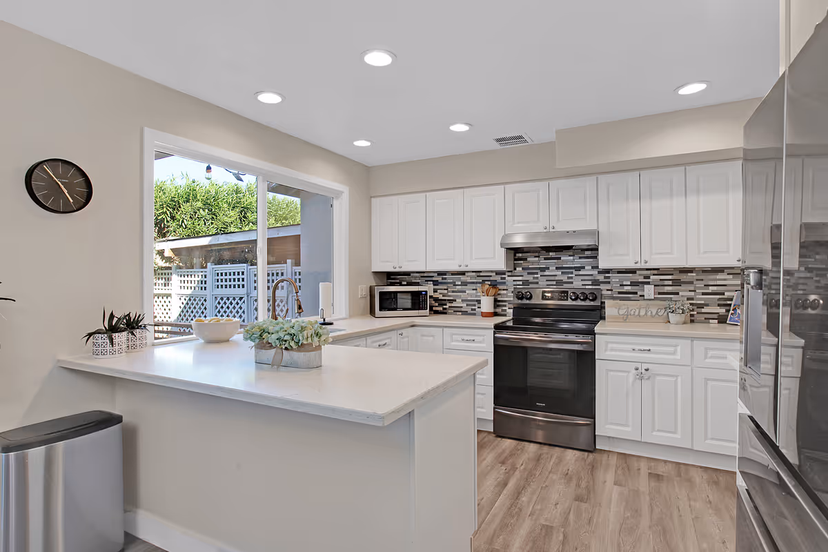 Bright modern kitchen with white cabinets, a tiled backsplash, stainless steel appliances including a stove and refrigerator, a large window overlooking an outdoor area, and a white countertop island with decorative plants.