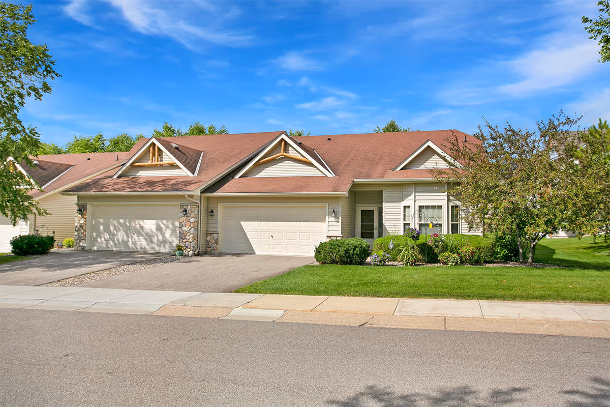 Two-unit beige residential building with attached double garages, a manicured front lawn and trees under a blue sky.