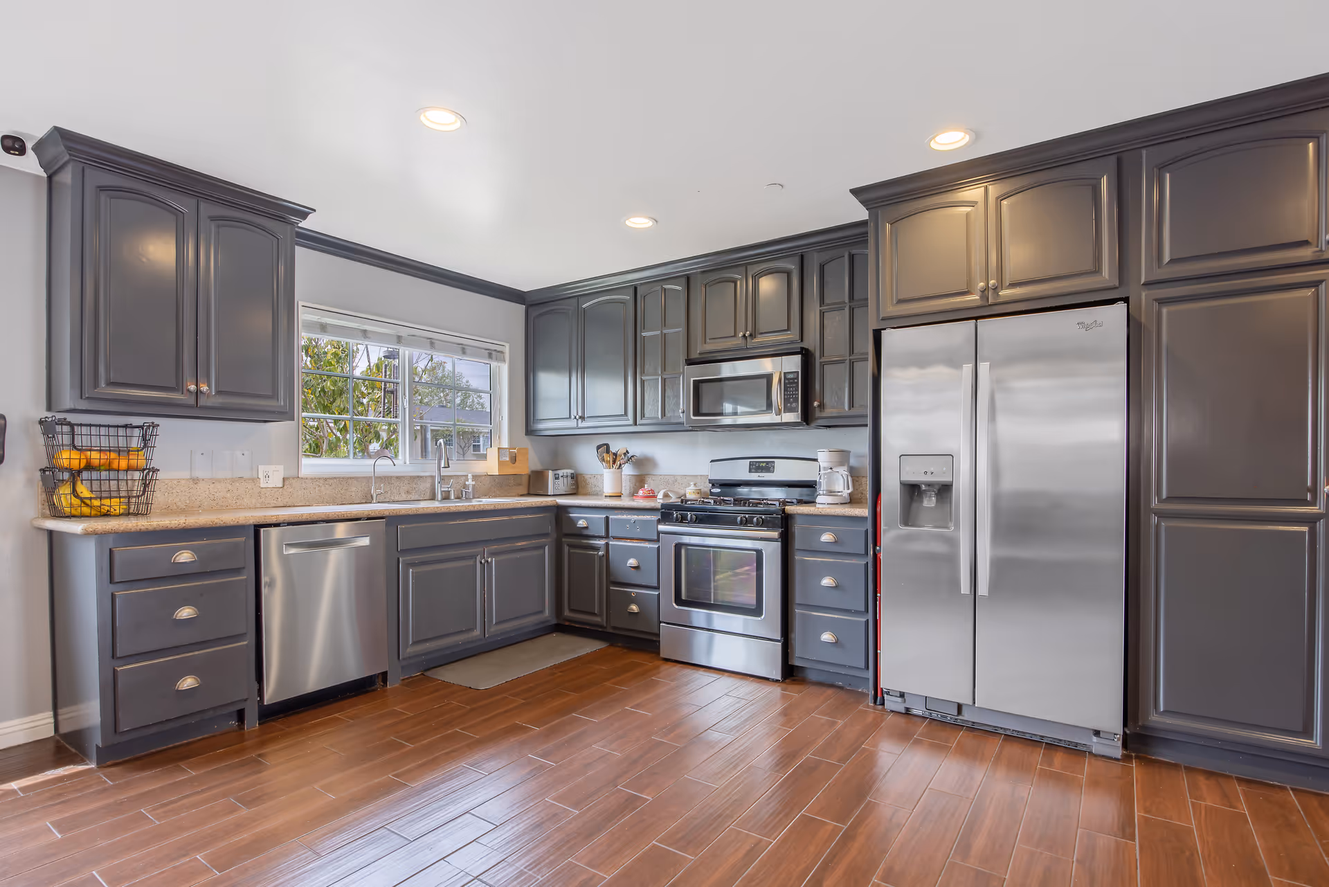 Modern kitchen with dark gray cabinets, stainless steel appliances including a refrigerator, oven, microwave, and dishwasher, a window above the sink, and wooden floor tiles.