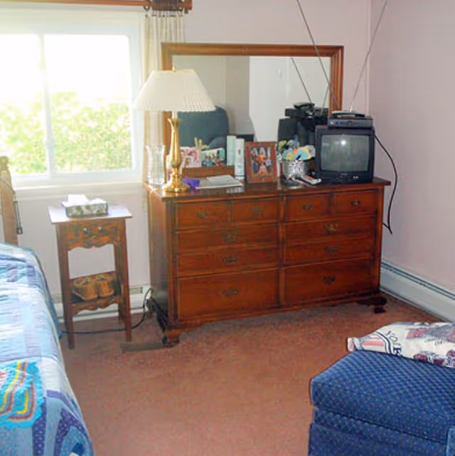 A bedroom with a wooden dresser topped with a lamp, framed photos, and a small television. There is a window with curtains letting in natural light, a small wooden side table with a tissue box, part of a bed with a colorful quilt, and a blue upholstered chair with a patterned pillow.