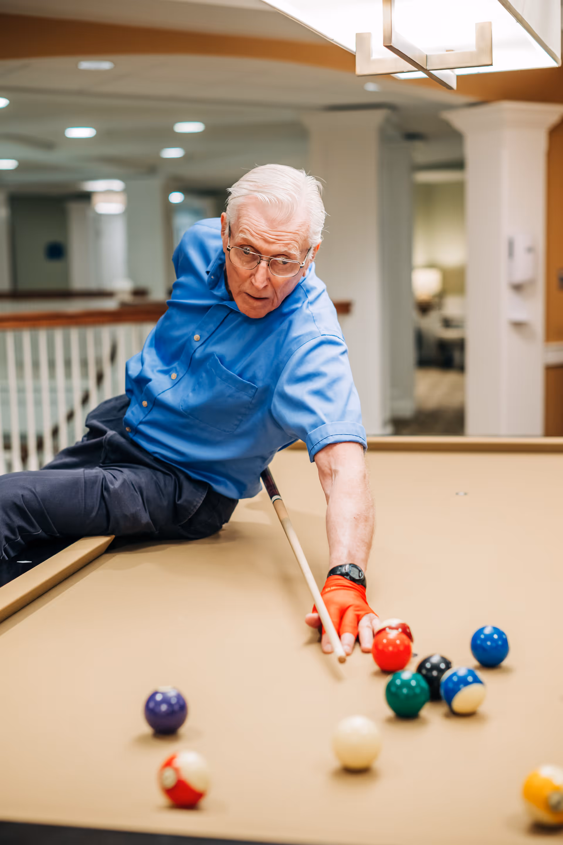 An elderly man wearing a blue shirt and an orange glove is playing billiards indoors, leaning over the pool table to take a shot with a cue stick.