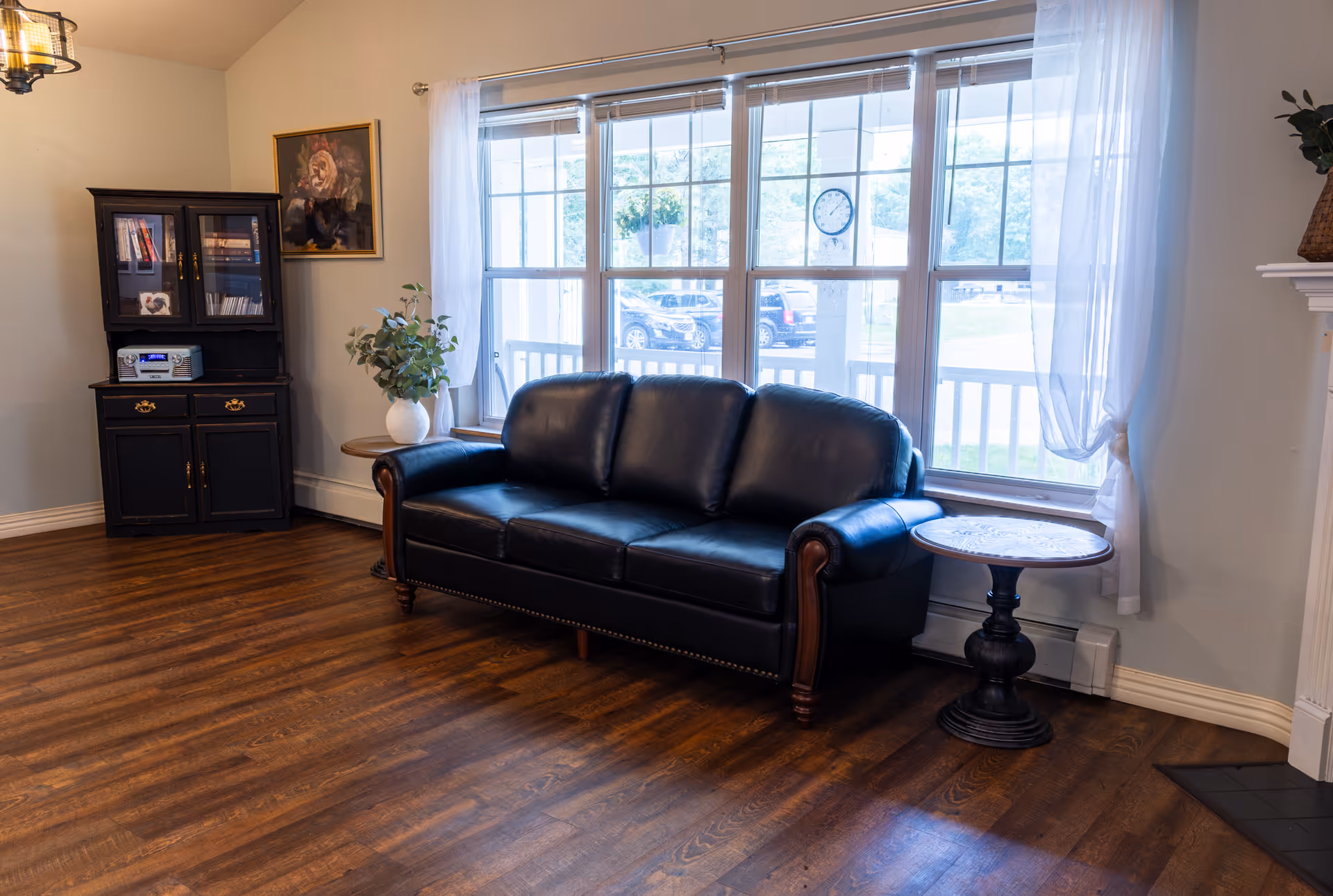 A cozy living room area with a black leather sofa positioned in front of large windows with sheer white curtains. To the left of the sofa is a small round wooden side table with a white vase holding green foliage. Further left is a black cabinet with glass doors containing books and a stereo system. The room has wooden flooring and light-colored walls, with a framed floral painting hanging above the cabinet.