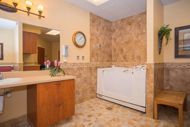 A clean and well-lit bathroom with beige and brown tiled walls and floor. The room features a white walk-in bathtub with safety handles, a wooden vanity with a sink and a mirror above it, a wall-mounted clock, a soap dispenser, a wooden bench, a wall-mounted plant, and a framed picture.