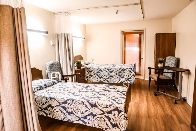 A senior living bedroom with two twin beds covered in patterned blue-and-white bedspreads, chairs, a nightstand, and wood-look flooring.