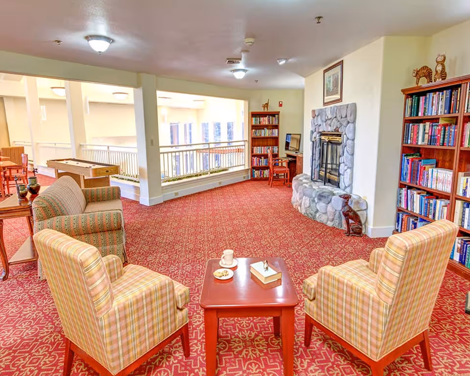 A cozy common area in a retirement and assisted living facility featuring two plaid armchairs and a small red table with a cup, plate of cookies, and tissue box. The room has a red patterned carpet, a stone fireplace with a dog statue, two wooden bookshelves filled with books, and a computer desk in the background. There is also a shuffleboard table and additional seating near a railing overlooking a lower level.