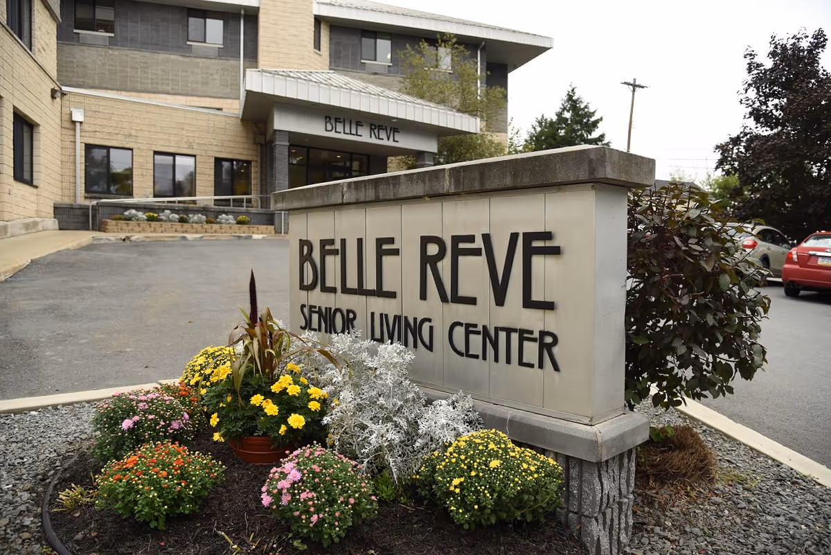 Stone sign reading "Belle Reve Senior Living Center" with flowers in front and the facility entrance and parking lot behind it.