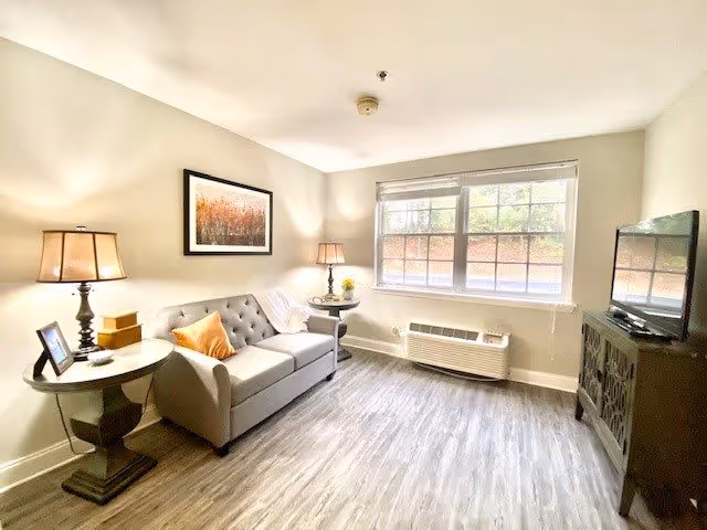 A bright living room with a gray tufted sofa adorned with an orange pillow and a white throw blanket. There are two side tables with lamps on either side of the sofa. A framed picture hangs on the wall above the sofa. A large window with blinds lets in natural light, and below it is a wall-mounted air conditioning unit. Across from the sofa is a dark wooden cabinet with a flat-screen TV on top. The floor is covered with light wood laminate.
