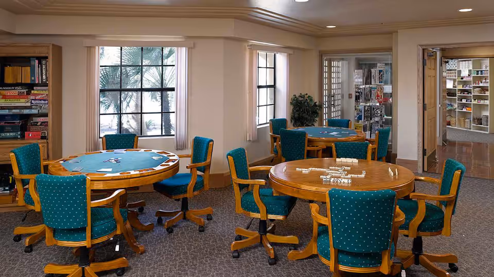 A well-lit game room with three round wooden tables, each surrounded by green upholstered chairs on wheels. The tables have game pieces on them, including dominoes and cards. Large windows with white curtains let in natural light, and there are bookshelves filled with board games and magazines in the background.