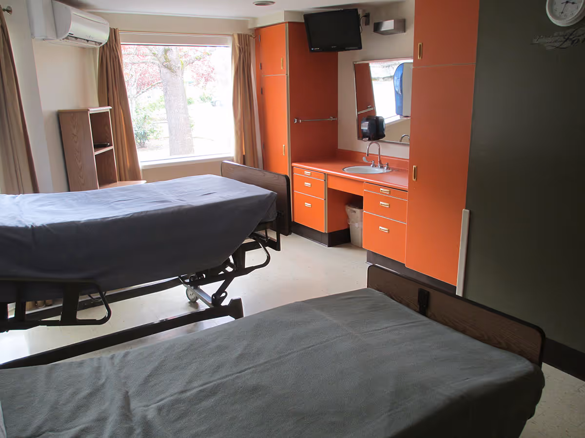 A nursing facility patient room with two hospital beds, orange built-in sink cabinets, a wall-mounted TV, and a large window.