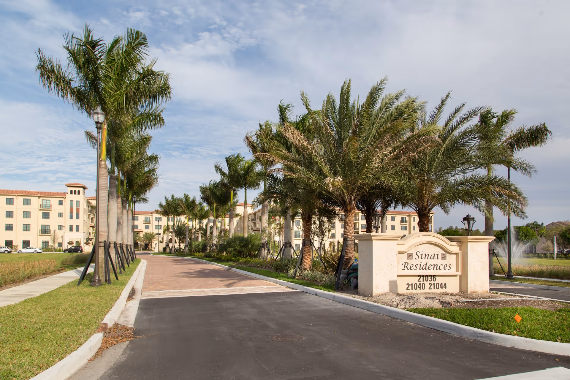 Entrance driveway lined with palm trees leading to a large multi-story building with beige walls and red-tiled roof. A stone sign at the entrance reads 'Sinai Residences' with address numbers below. There is a fountain visible in the background on the right side.